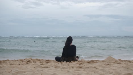 rear view of young, woman sitting in deep thought at white sand beach with strong winds. -static shot