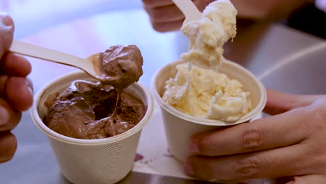 a hand uses a wooden spoon to scoop chocolate and vanilla ice cream into paper cups on a reflective table under soft, natural lighting. close-up perspective