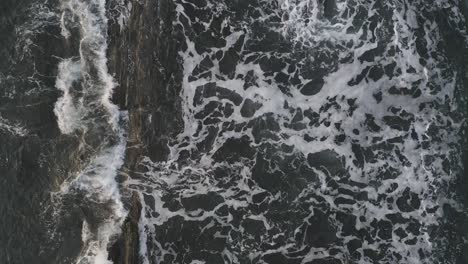 flying slowly forward as waves crash on rocky shore at sunrise aerial top down