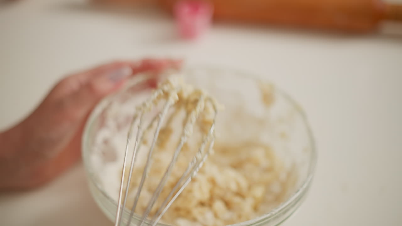 close up of fair skinned person holding metal whisk covered with dough, tapping it on glass bowl to release sticky mixture, small splash lands on hand and is returned to bowl during baking process