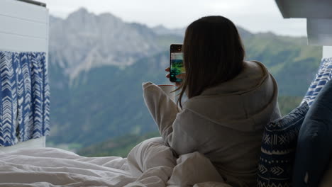 Young-woman-hold-mug-of-coffee-and-taking-pictures-with-smartphone-of-Dolomites-from-bed-on-travel-van