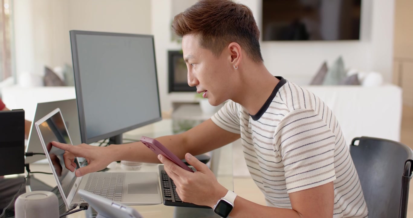 Uomo asiatico di mezza età che usa un telefono rosa come riferimento, digita e tocca lo schermo del laptop gestendo il lavoro.