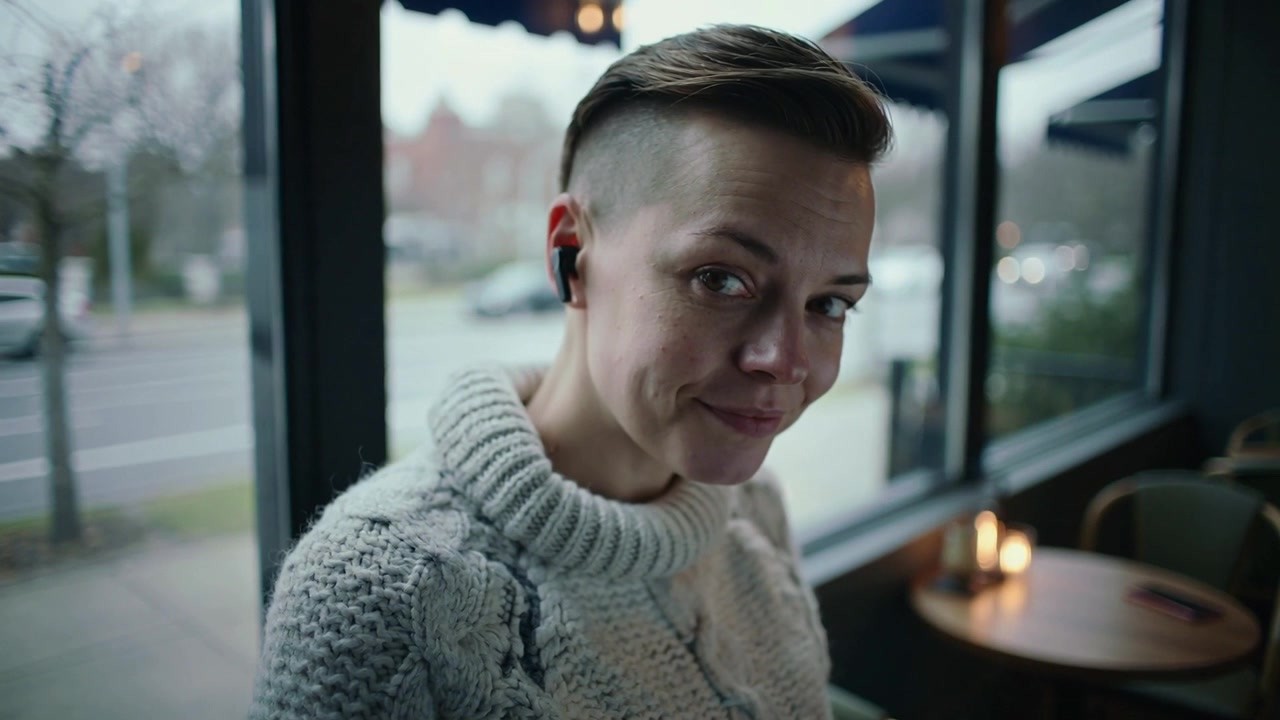 Noticing camera, woman in chunky turtleneck and earbuds turning to camera at cafe table, candle lit