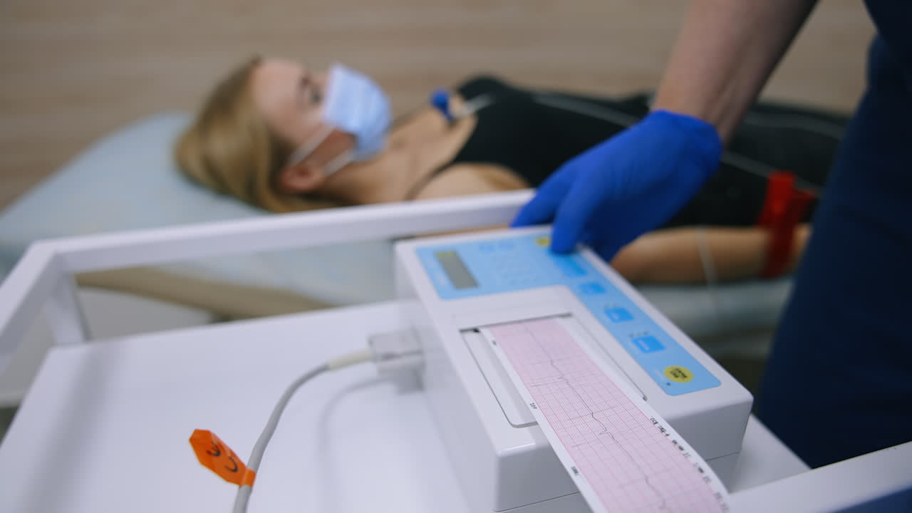 Paper with heart test result appears from electrocardiograph. Close up. Medic's gloved hand tears the paper from machine. Female patient at backdrop in blur.