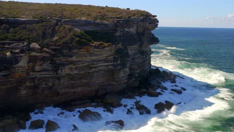 Olas-Rompiendo-Sobre-Rocas,-Movimiento-De-Agua-De-Surf-En-Acantilados-Escarpados-En-El-Parque-Nacional-Real-En-Nueva-Gales-Del-Sur,-Australia---Toma-Aérea