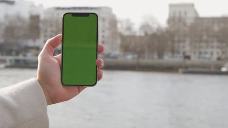 primer plano de un hombre que sostiene un teléfono móvil de pantalla verde con vistas al río támesis y al terraplén en londres, reino unido