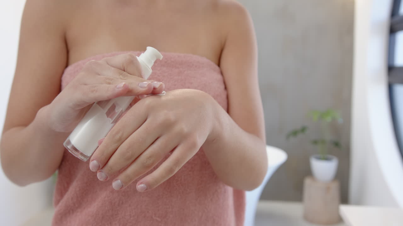 Applying lotion, woman in towel focusing on skincare routine in bathroom
