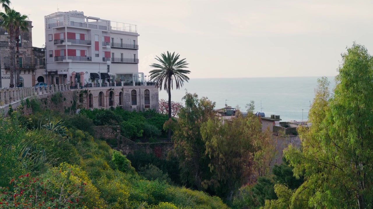 Un edificio storico con balconi con persiane rosse e un'alta palma si erge sul mare, catturando il fascino costiero della Sicilia