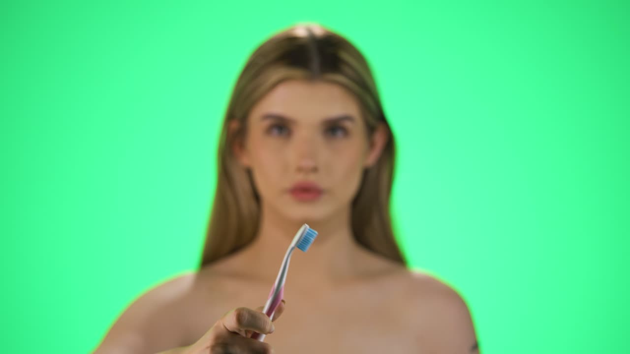 Medium shot of a pretty young woman with a toothbrush in her hand holding it up to the camera and starting to brush her teeth for good dental hygiene and healthy teeth in front of green background