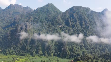 aerial flying past floating clouds with forested mountains of vang vieng in background