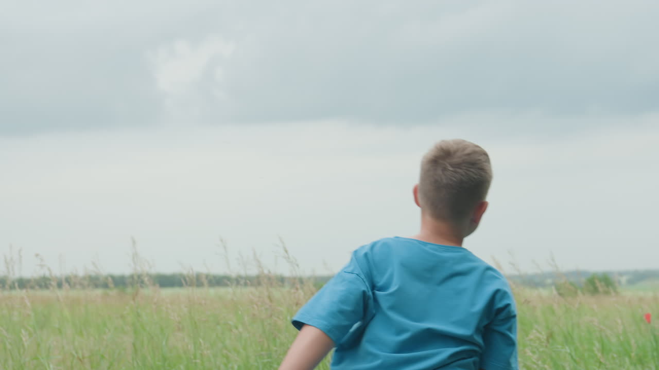 푸른색 셔츠와 반바지를 입고 라켓을 든 백인 소년이 거친 초원을 뛰어다니며 키 큰 풀밭을 활기차게 뛰어넘고, 흐릿한 수평선, 활발한 추격전과 장난기 넘치는 질주, 활동적인 여름 야외 풍경