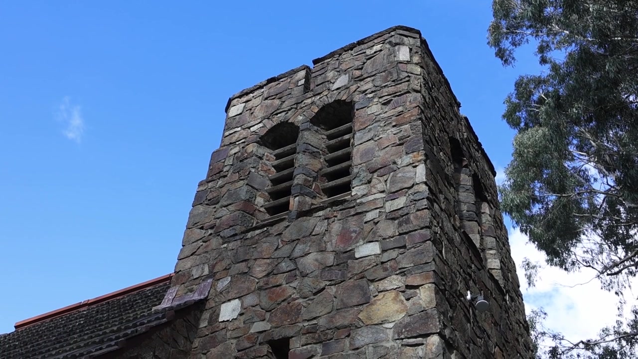 Robusta torre in pietra di una vecchia chiesa a Olinda, Melbourne, ripresa con una panoramica costante verso l'alto contro un cielo blu vibrante e alberi di eucalipto, senza persone nell'inquadratura. Perfetto per stock architettonico.