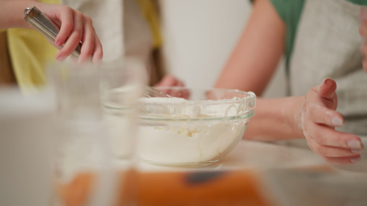 close up of metal whisk lifting sticky flour and egg dough mixture from transparent bowl while person with flour-stained hands sits nearby in kitchen setting with blurred foreground objects