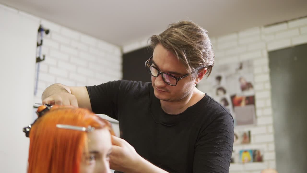 Stylish male hairdresser in glasses is cutting redhead woman's hair in beauty salon. Young woman getting her haircut and