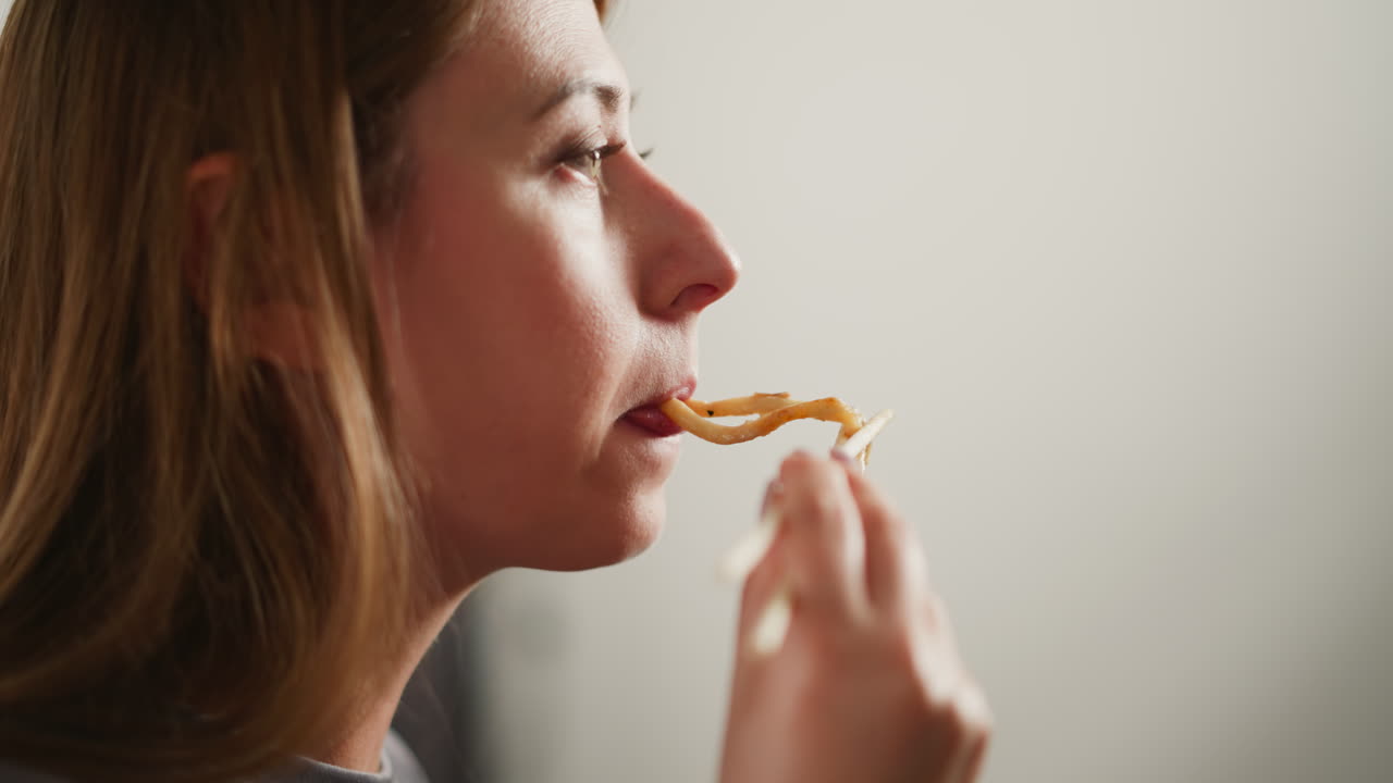 Close up side view of woman eating noodles using chopsticks, standing in bright modern kitchen with soft blurred white background, focusing on relaxed expression