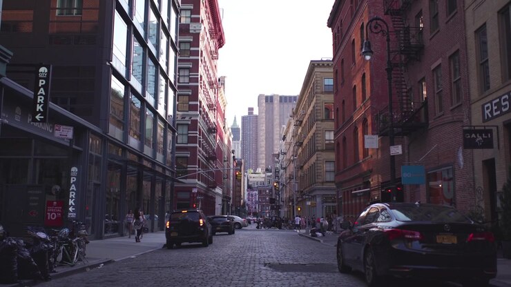 NYC: Mercer Street, SoHo, rivolta a sud verso il Woolworth Building in lontananza, con pedoni, auto e attività commerciali in primo piano, immersa nell'ombra e nella luce - New York City, USA