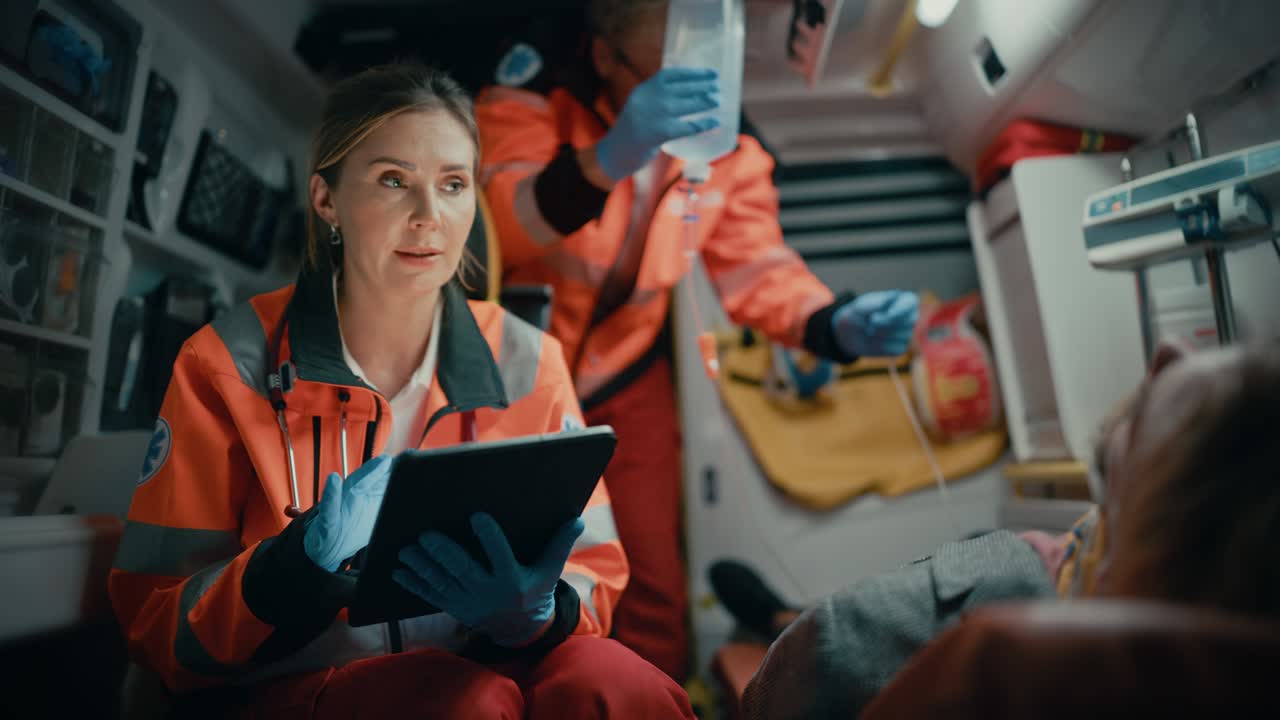 профессиональная фельдшерка использует планшетный компьютер, чтобы заполнить анкету для раненого пациента по пути в больницу. помощник скорой помощи утешает пациента в скорой помощи.