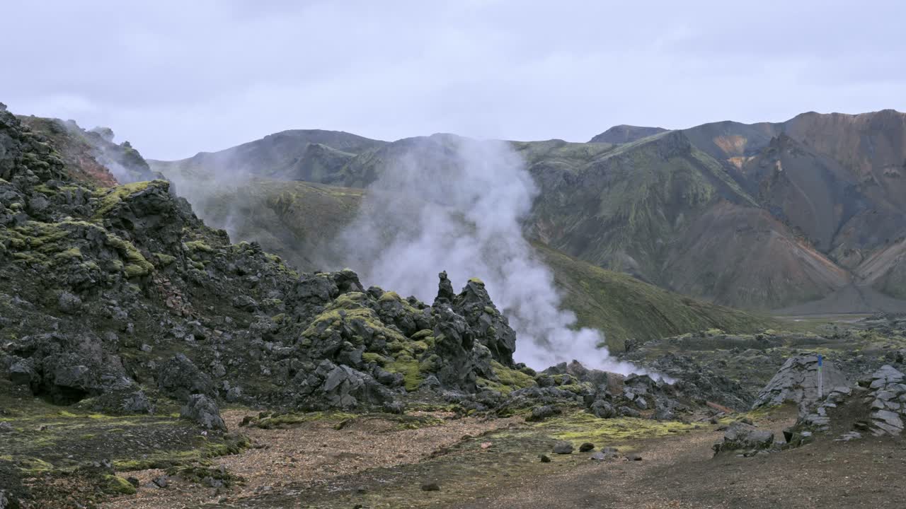 Uap mengepul dari mata air panas bumi di antara bebatuan lava hitam di lanskap vulkanik Landmannalaugar, Islandia yang berwarna-warni. Bidikan genggam