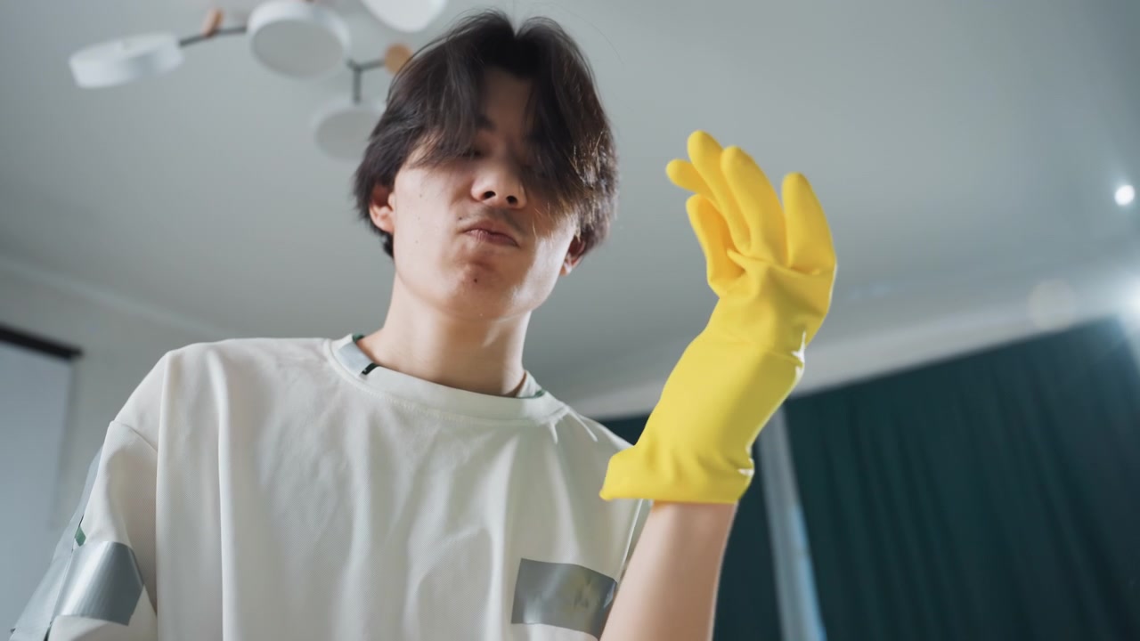 노란색 고무장갑을 낀 젊은 아시아 남성이 집중된 표정으로 앞으로 손을 뻗는 로우 앵글 클로즈업, 배경에는 천장 선풍기와 커튼, 보호 장갑을 낀 손으로 딥 클리닝을 준비하는 모습, 가정 위생