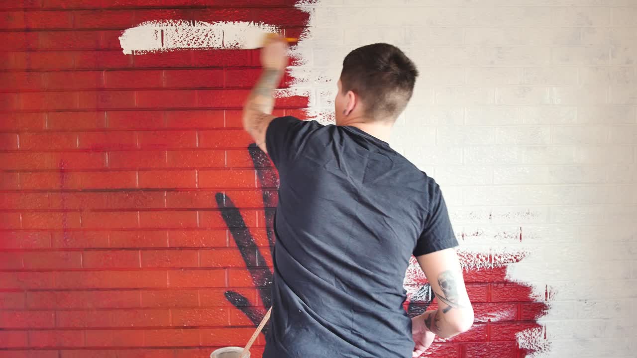 artista tatuado pintando sobre un mural rojo en una pared de ladrillo con pintura blanca