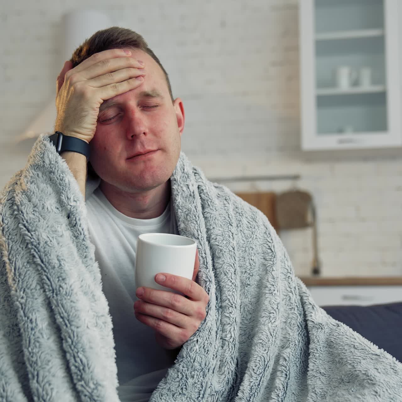 Unhappy guy suffering coronavirus infection. Sick man covered with warm blanket with a cup of hot beverage staying home. Self-isolation and quarantine