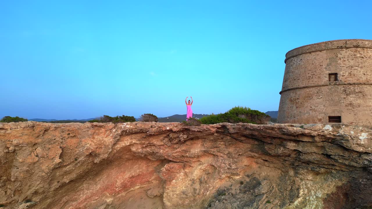 Carefree woman in pink dress with open arms enjoying freedom and breathtaking view on Ibiza island near Torre des Savinar at sunset. Majestic aerial view flight descending drone