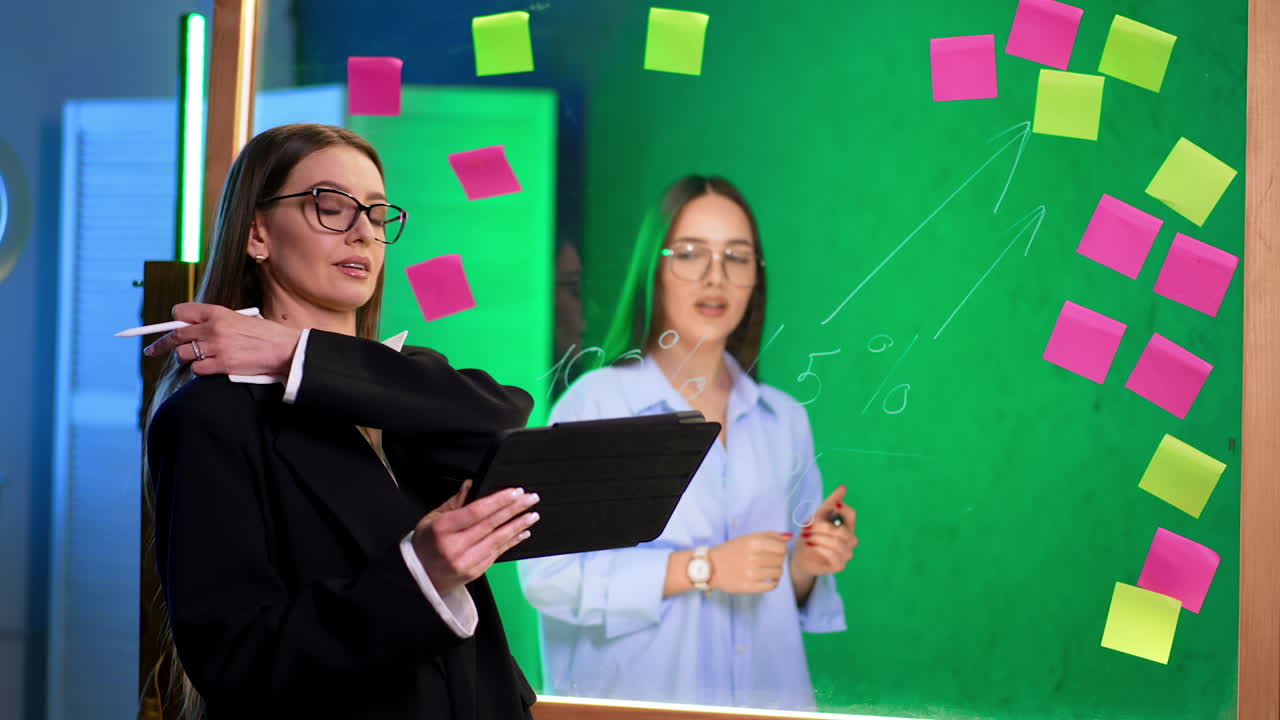 Two Caucasian ladies stand on both sides of glass wall. Woman standing at foreground holds the tablet. Girl behind the board writes with a marker.