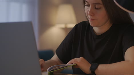 female-student-is-learning-online-sitting-at-home-at-evening-and-making-notes-of-webinar-and-lecture-viewing-on-laptop-portrait-in-living-room