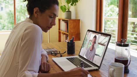 mujer afroamericana tomando notas mientras tiene una videollamada en una computadora portátil en casa