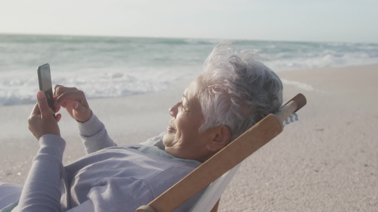 Happy hispanic senior woman relaxing on sunbed on beach at sunset, using smartphone