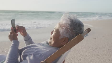 happy hispanic senior woman relaxing on sunbed on beach at sunset, using smartphone