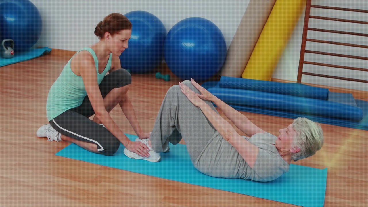 Anleitender Fitnesstrainer stabilisiert Senior in grauem T-Shirt im Fitnessstudio, der Crunches auf einer blauen Matte macht.