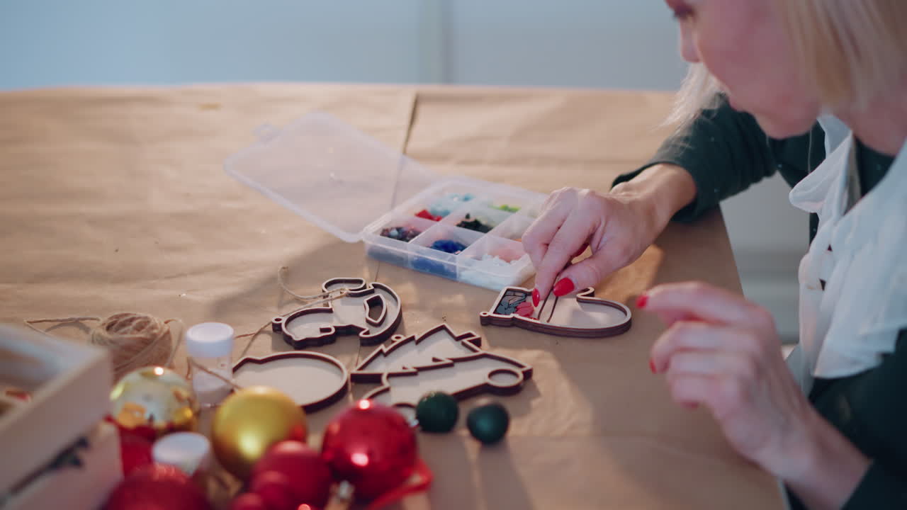 Donna che decora ornamenti natalizi in legno, accogliente laboratorio da tavolo, vassoio per colori, perline, palline di Natale, spago, nastro, mani in primo piano che assemblano ornamenti, preparazione di regali natalizi, luce naturale calda