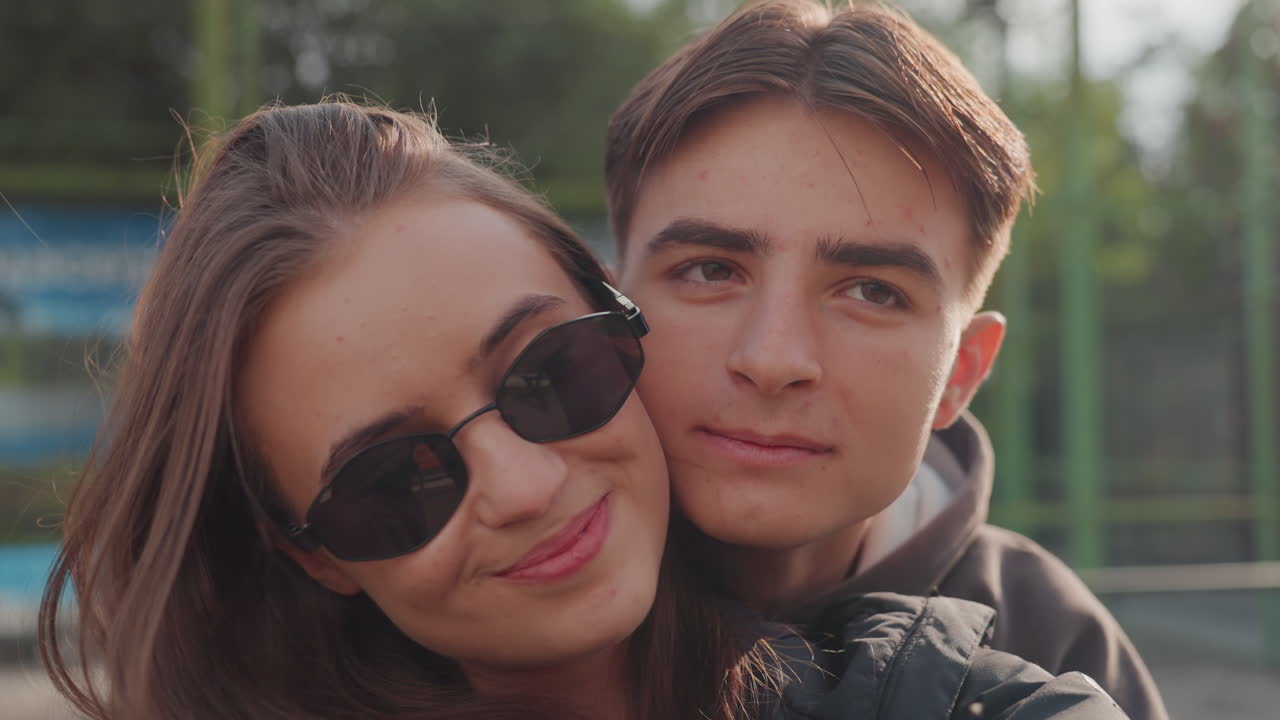 giovane coppia caucasica si scambia un bacio all'aperto, scena di appuntamento intima con sorrisi dolci e bacio sulla guancia in un parco suburbano. Uno smartphone portatile cattura primi piani intimi, luce solare calda e atmosfera nostalgica