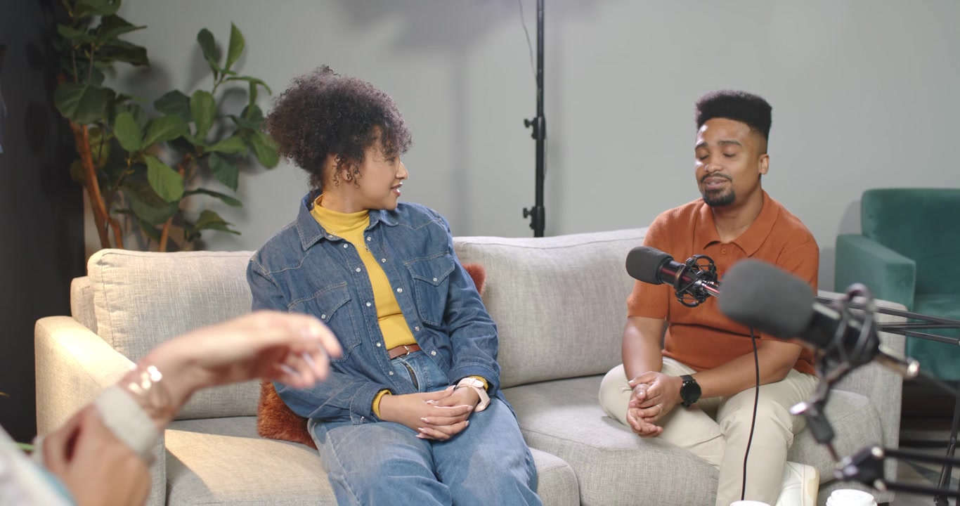Afroamerikanska podcastgäster svarar värden, man talar i soffan, kvinna i jeans, bom-mikrofon.