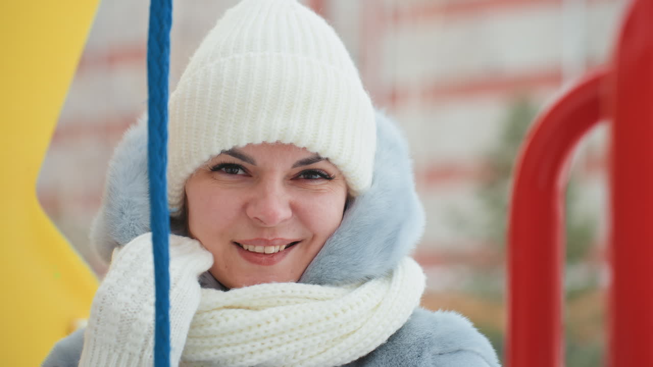 Mãe agasalhada em casaco de inverno no parquinho, sorrindo atrás dos corrimãos coloridos do escorregador. Usando gorro de malha e cachecol grosso, com bochechas rosadas do frio. Retrato espontâneo que transmite um calor materno aconchegante.