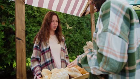 over the shoulder, a happy brunette guy in a green checkered shirt talks with a girl who sells baked goods in her shop during a fair against the backdrop of green coniferous trees in summer