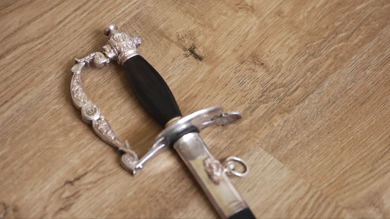 espada de plata detallada con diseños ornamentados y un mango negro, descansando en un piso de madera