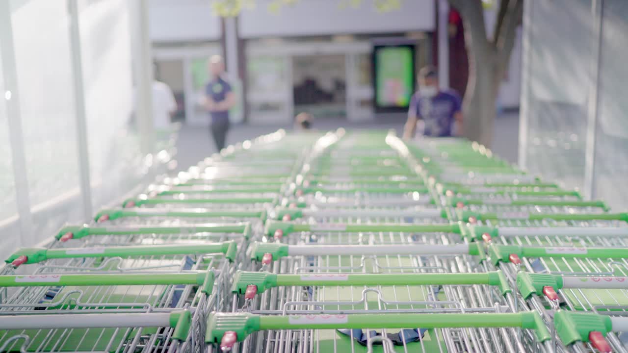 Shopping carts with green handles are stacked in row in front of a supermarket  entrance and some people walking in the background during a sunny day