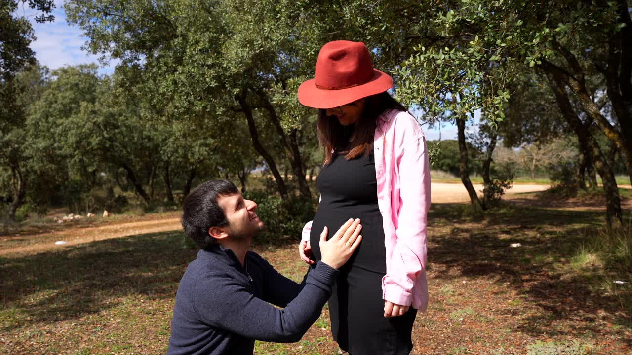 Smiling husband gently touches pregnant wife's belly while kneeling as she strokes his hair outdoors