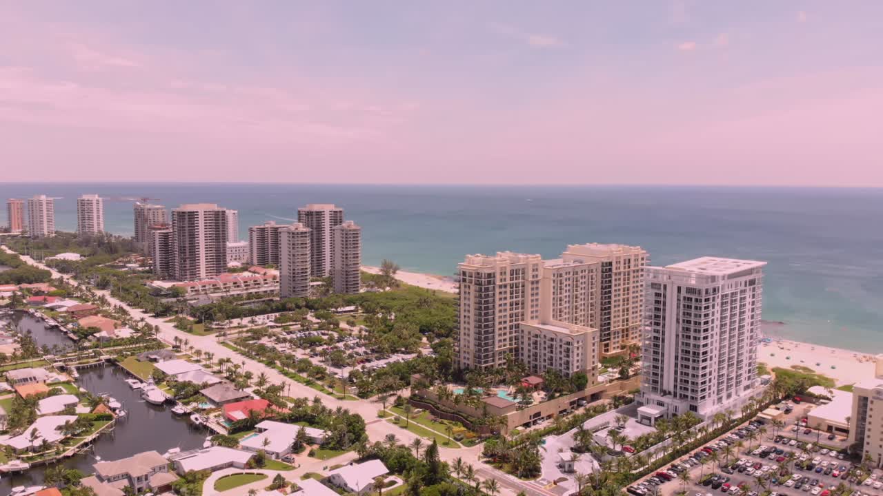 A drone shot flying backwards captures a beachfront hotel at sunset, revealing the sandy shores, ocean waves, and the hotel’s pool under a golden sky.