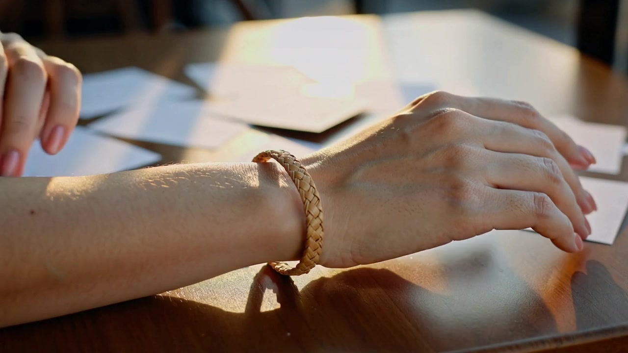 Reaching right hand, woman adjusting braided bracelet then resting left arm at sunlit table, cards