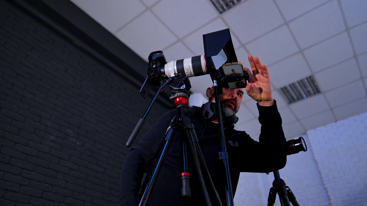 Cameraman che regola l'obiettivo della telecamera e la configurazione del telefono in studio. Inquadratura dal basso di un uomo con le cuffie che sistema l'attrezzatura professionale per la ripresa e un dispositivo mobile su treppiede.