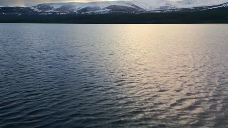 aerial drone footage begins overlooking a tranquil scottish lake, ascending to reveal a mesmerizing panorama of majestic snow-clad mountains and vast highland terrain