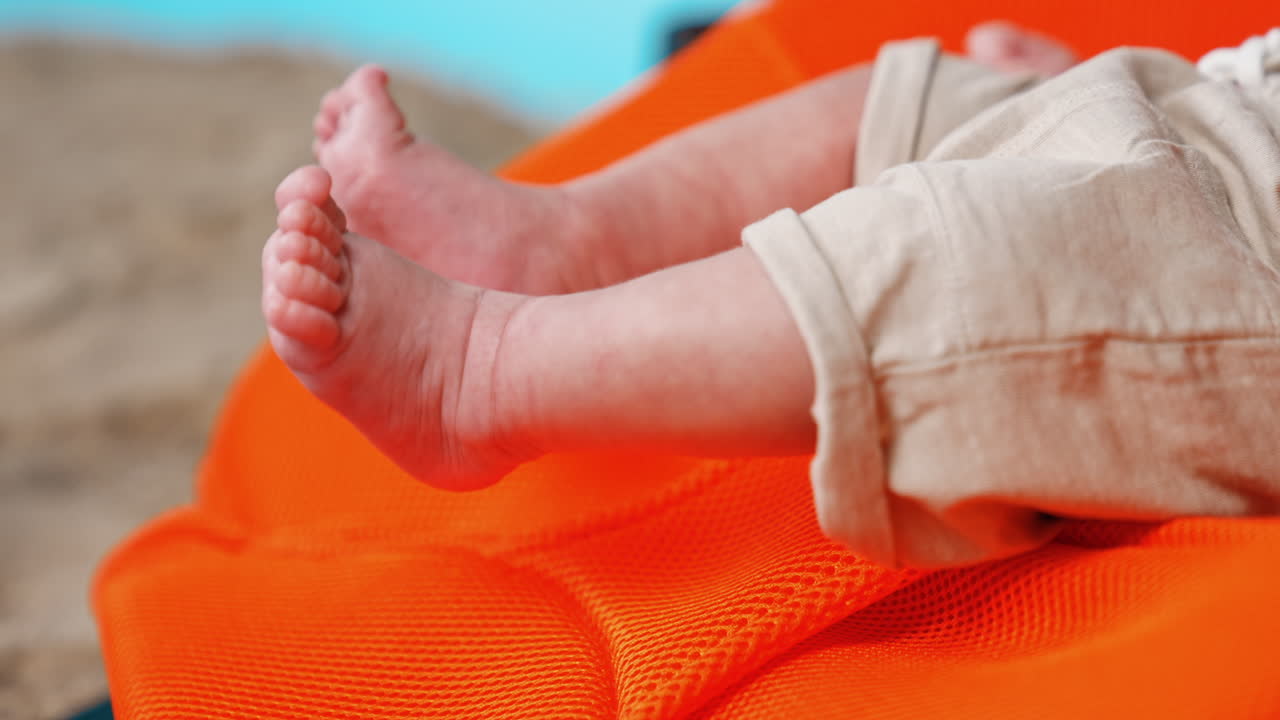 Cute tiny bare feet of a Caucasian baby lying in the orange chair. Newborn in shorts moving his legs. Close up.
