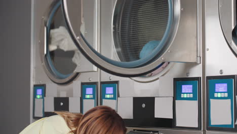 female worker focused on transferring pile of clothes from washer into dryer inside laundromat, multitasking while handling daily laundry operations with care and precision