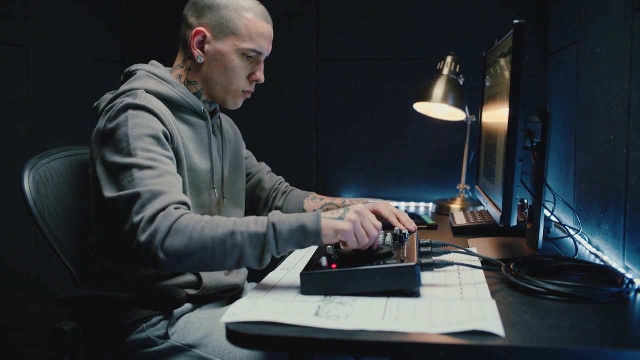 DJ tatuato in felpa grigia che regola il controller e mixa tracce alla scrivania, mentre la telecamera si allontana.