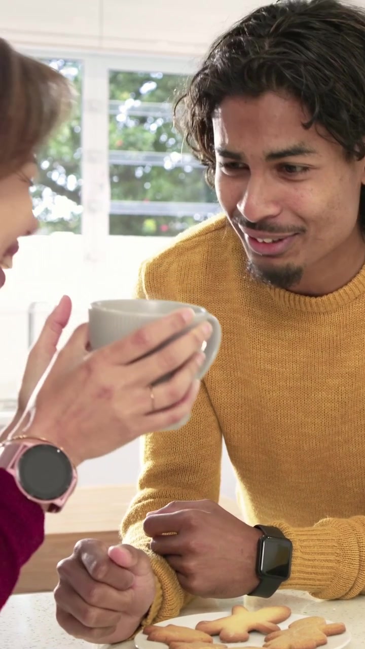 Vertical video: Offering mug, pair sharing cookies and chatting in kitchen wearing burgundy mustard