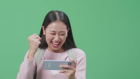 행복한 아시아 여성 학생이 모바일 게임을 하고 녹색 스크린 스튜디오에서 는 동안 승리합니다.
