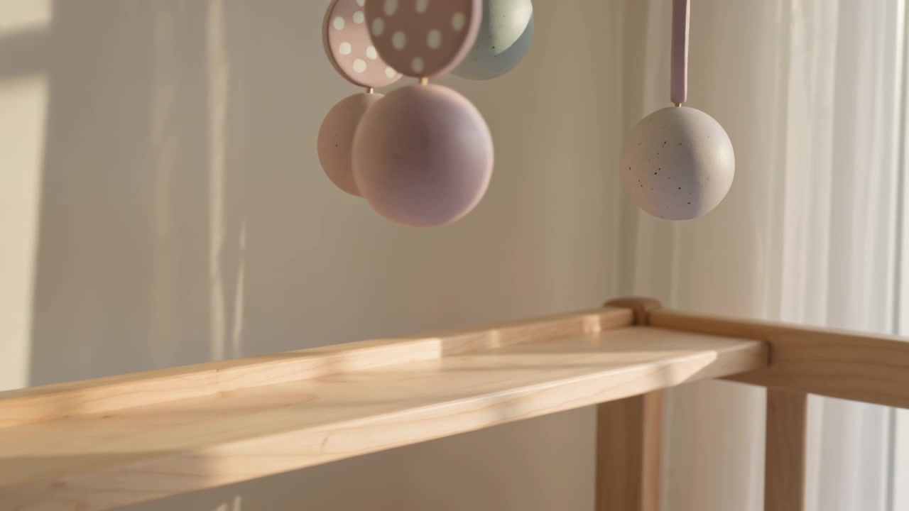 Swinging pastel mobile rotating above wood crib rail at window, sun streaming through sheer drape